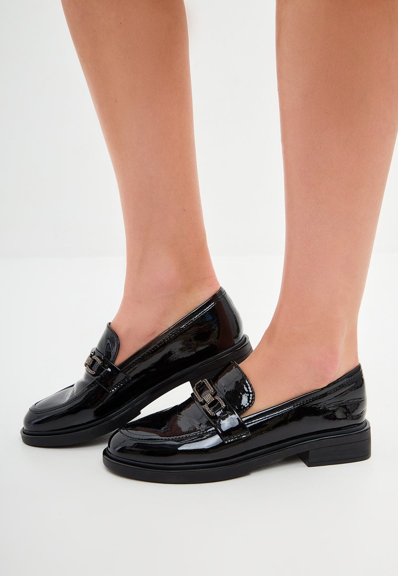 Black patent leather loafers with a low block heel, featuring a rounded toe and a decorative buckle detail on the upper.