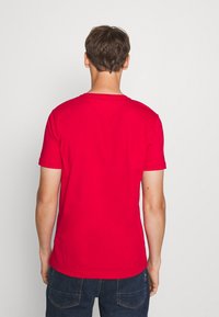 Joven de espaldas, vistiendo una camiseta roja de manga corta y jeans azul oscuro, sobre un fondo blanco.