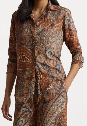 Femme portant une chemise à manches longues et un pantalon marron assortis à motif cachemire, une main dans la poche et cheveux foncés lâchés.