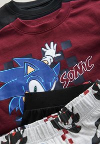 Mikina v červené a černé barvě se Sonic motivem a grafickým designem, zobrazující modrou postavu. Dole černé šortky s bílým vzorem.