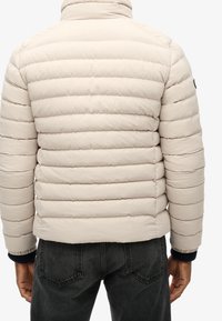Uomo che indossa un giubbotto trapuntato beige chiaro e jeans grigio scuro, visto da dietro su uno sfondo bianco.