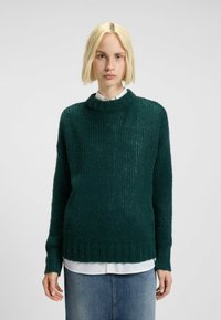 Maglione verde a maglia con una vestibilità ampia, texture a costine e collo alto. Indossato sopra una camicia bianca, presenta maniche lunghe e una silhouette rilassata.