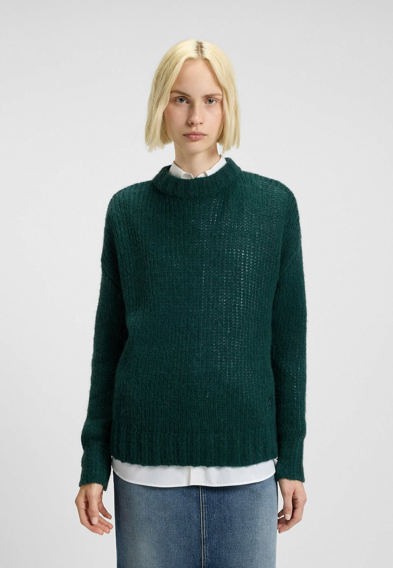 Maglione verde a maglia con una vestibilità ampia, texture a costine e collo alto. Indossato sopra una camicia bianca, presenta maniche lunghe e una silhouette rilassata.