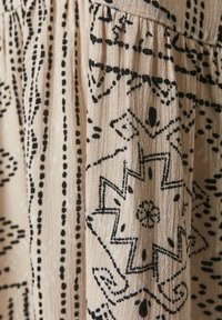 Beige tyg med ett intrikat svart mönster av linjer och geometriska former, med en texturerad yta och draperade designdetaljer.