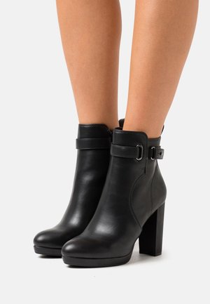 Paire de bottines en cuir noir avec talons carrés hauts et détail de boucle à sangle, portées sur des jambes nues, sur fond blanc.