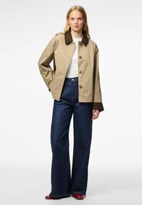 Mujer rubia vestida con una chaqueta beige con cuello y puños oscuros, camisa blanca, jeans de pierna ancha de color azul oscuro y zapatos rojos, de pie contra un fondo blanco.