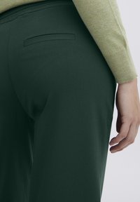 Green trousers with a smooth texture and tailored design. Features a narrow back pocket and a fitted waistband, paired with a light sweater.
