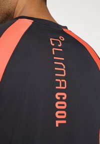 Cămașă sport neagră cu accente din plasă roșie, gât rotund și textul 'CLIMA COOL' imprimat vertical cu bold în portocaliu pe spate.