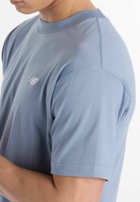 T-shirt bleu clair à manches courtes avec un col rond, texture lisse et un petit logo blanc sur la poitrine. Présenté porté par une personne.