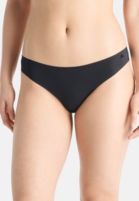 Frau in schwarzer Bikini-Unterwäsche steht vor einem schlichten Hintergrund und zeigt den Oberkörper und die Oberschenkel.