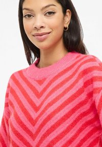 Pull rayé rose et rouge avec un motif chevron, col rond côtelé, texture douce et coupe décontractée.