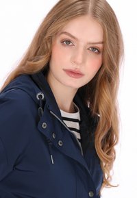 Granatowa wodoodporna kurtka z wysokim kołnierzem, regulowanym kapturem ze ściągaczem oraz srebrnymi elementami. Pod spodem widoczna koszula w paski.