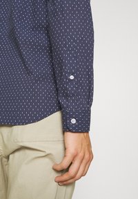 Gros plan d'une personne portant une chemise bleu marine à manches longues avec de petits motifs blancs en tirets et un pantalon beige, montrant le poignet et les boutons.