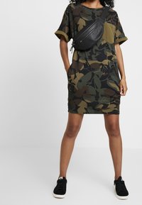 V-aukkoisen, lyhythihaisen camo-kuosiset T-paitamekko vihreänä ja ruskeana, joissa on sivutaskut, sekä musta vyölaukku kuljetettuna vinosti rinnan yli.