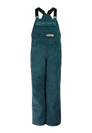 Junge Person trägt türkisfarbene Ski-Overalls, ein schwarzes langärmeliges Oberteil mit Sternenmuster, schwarze Stiefel, schwarze Ohrenschützer und schwarze Sonnenbrille.