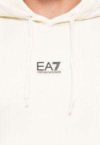 Hvit hettegenser med snøring som har svart "EA7 Emporio Armani"-logo sentrert på brystet.