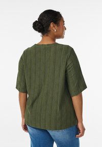 Zelený texturovaný top s krátkými rukávy, kulatým výstřihem a dekorativními vzory. Nošený s modrými džínami, pohled zezadu.