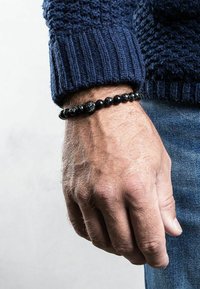 Musthaveehelmed mustavast helmed pärlitega käevõru, millel on siledad ja tekstureeritud pärlid, kantud heleda nahaga käel, koos sinise sviitri ja teksadega.