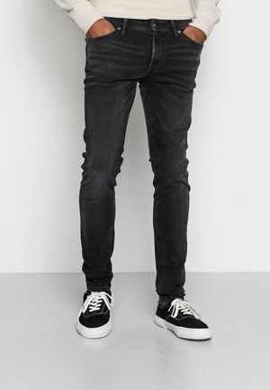 Jeans pretos em corte slim feitos de denim com uma textura ligeiramente desbotada, apresentando bolsos frontais e usados com ténis pretos.
