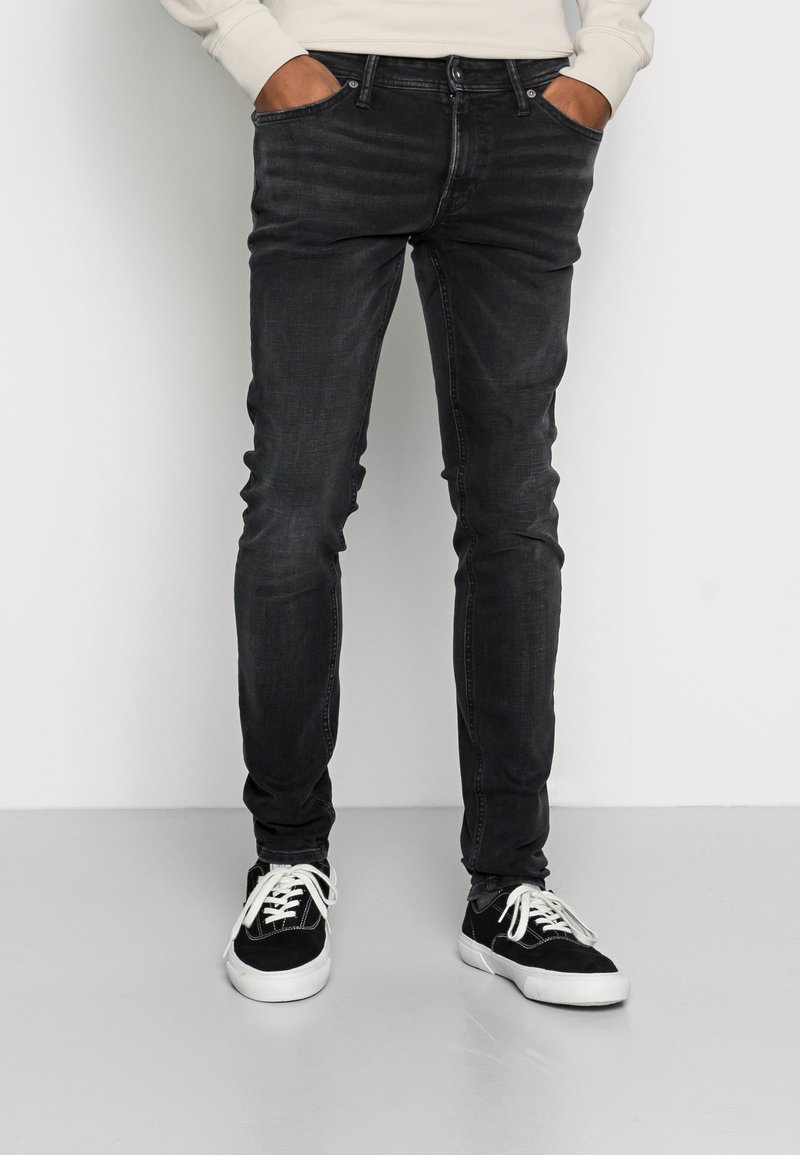 Jeans pretos em corte slim feitos de denim com uma textura ligeiramente desbotada, apresentando bolsos frontais e usados com ténis pretos.