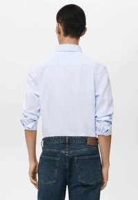 Camicia a righe blu chiaro con colletto piegato e maniche arrotolate, abbinata a jeans in denim scuro con una toppa in pelle marrone e tasche posteriori.