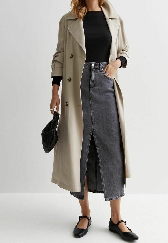 Femme portant un trench-coat beige par-dessus un haut noir et une jupe en denim gris fendue à l'avant, tenant un sac à main noir et portant des ballerines noires.