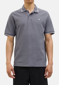 Polo gris en tissu texturé, avec un liseré blanc le long du col et des manches, et un petit logo sur la poitrine. Manches courtes, coupe classique.