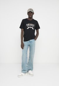 Crna majica s bijelim natpisom "ARCHIVE", ležernog kroja, uparena s svijetloplavim trapericama na zvono i bež tenisicama. Model nosi bež kapu.