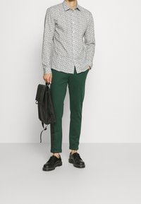 Blommönstrad skjorta med knäppning i vitt och svart, ihopparad med mörkgröna smala byxor och svarta snörskor, hållande en svart ryggsäck.