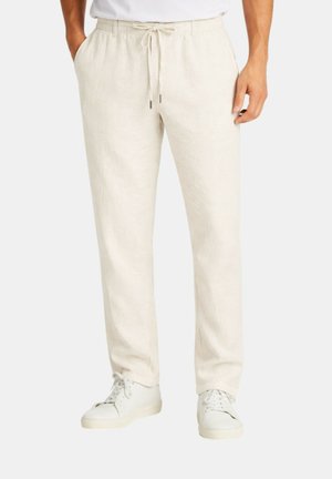 Uomo che indossa pantaloni di lino color avorio con coulisse e sneakers bianche, in piedi con una mano in tasca su sfondo semplice.