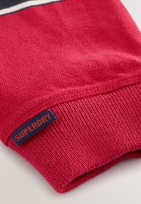 Puño de suéter de punto rojo con textura acanalada, con una etiqueta cosida de color azul marino que muestra "SUPERDRY" en letras naranjas contrastantes.