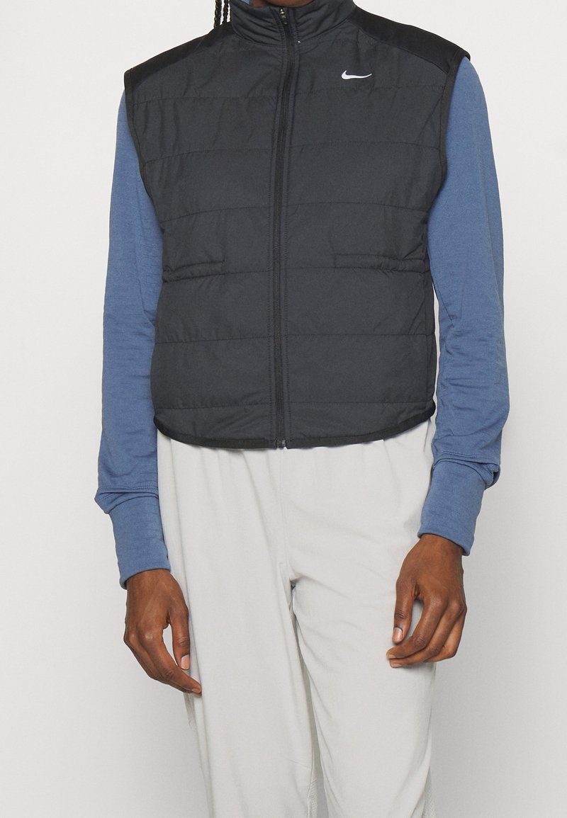 Personne portant un gilet matelassé sans manches noir Nike par-dessus une chemise bleue à manches longues et un pantalon gris clair, debout, les bras détendus.