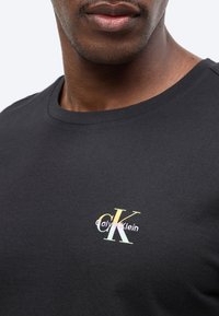 Gros plan d'une personne portant un T-shirt noir avec un petit logo Calvin Klein pastel sur la poitrine.