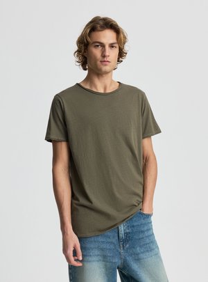 Giovane uomo con capelli biondi ricci che indossa una maglietta verde oliva e jeans blu, in piedi davanti a uno sfondo grigio chiaro uniforme.