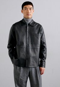 Svart läderjacka med krage, framknappar och en ficka med patch. Slät textur, avslappnad passform, kombinerad med grå byxor.