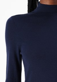 Haut en tricot bleu marine à manches longues avec un col montant. Texture lisse avec une coupe cintrée, mettant en valeur les épaules et les bras.
