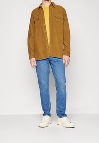 Brun manchester skjorta, gul t-shirt under, blå jeans och vita sneakers. Knapp-up design med bröstfickor. Klassisk passform.