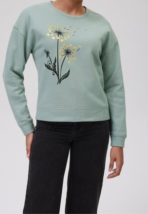 Personne portant un sweat-shirt vert sauge avec un imprimé pissenlit noir et doré, associé à un jean noir taille haute.