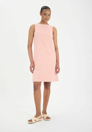 Robe sans manches rose avec une coupe droite et un tissu texturé, associée à des sandales beige clair, se tenant sur un fond neutre.