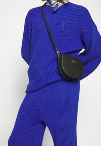 Ensemble pull en maille et pantalon en bleu vif, avec des côtes texturées et des détails découpés. Sac bandoulière noir avec accent logo.
