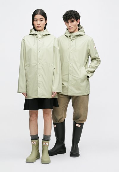 Δύο μοντέλα φορούν ταιριαστά ανοιχτόχρωμα πράσινα αδιάβροχα μπουφάν με κουκούλες, γκρι κάλτσες και μπότες Hunter. Η μία φοράει μαύρες μπότες και παντελόνι, ενώ η άλλη έχει πράσινες μπότες και φούστα.