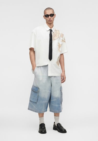 Νέος άνδρας που φορά γυαλιά ηλίου, λευκό πουκάμισο με κοντά μανίκια και floral σχέδιο, μαύρη γραβάτα, φαρδιά ξεβαμμένα τζιν σορτς με τσέπες, μπεζ κάλτσες και μαύρα μοκασίνια.