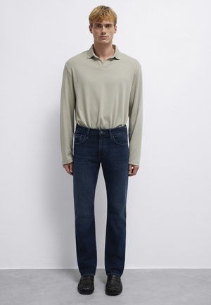 Blonder Mann steht, trägt ein hellbeiges langärmeliges Poloshirt, dunkelblaue Jeans und schwarze Schuhe vor einem weißen, einfarbigen Hintergrund.