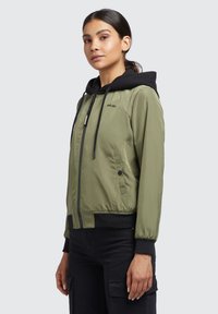 Jachetă bomber de culoare verde măslinie cu glugă neagră, fermoar frontal, buzunare laterale, manșete ribbate și detaliu cu logo pe piept. Material neted, ușor.