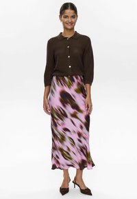 Cardigan marrone a coste, abbottonato al colletto. Gonna rosa fluida con motivi astratti verdi e marroni scuri. Tacchi a punta marroni.