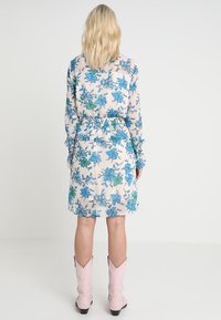 Femme blonde portant une robe rose pâle jusqu'aux genoux avec un imprimé floral bleu et vert, se tenant dos à la caméra, associée à des bottes rose pâle mi-mollet.