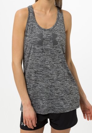 Kobieta nosząca szary sportowy top bez rękawów z czarnym logo Under Armour, w połączeniu z czarnymi sportowymi szortami, stojąca z rozluźnionymi ramionami.