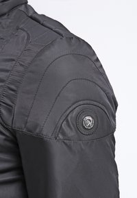 Svart vadderad jacka med en strukturerad axel, quiltade sömmar och en distinkt läderliknande lapp med detalj av knapp.