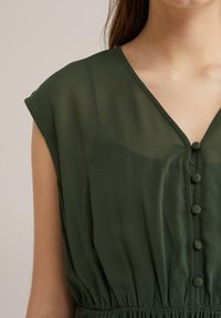 Zelené šaty s krátkým rukávem, výstřihem do V, knoflíkovou légou a elastickým pasem. Vyrobené z lehkého, průhledného materiálu.