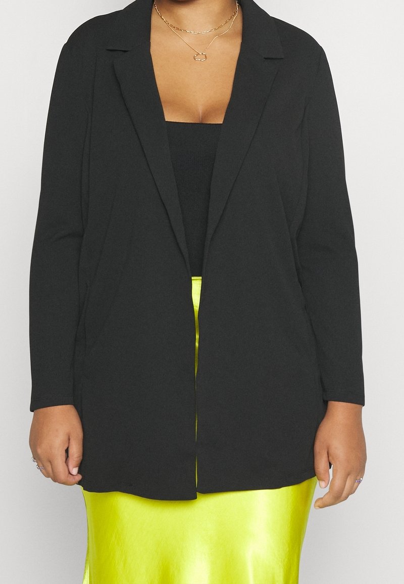 Personne portant un blazer noir ouvert devant, un haut noir, des colliers dorés superposés et une jupe brillante jaune vif.